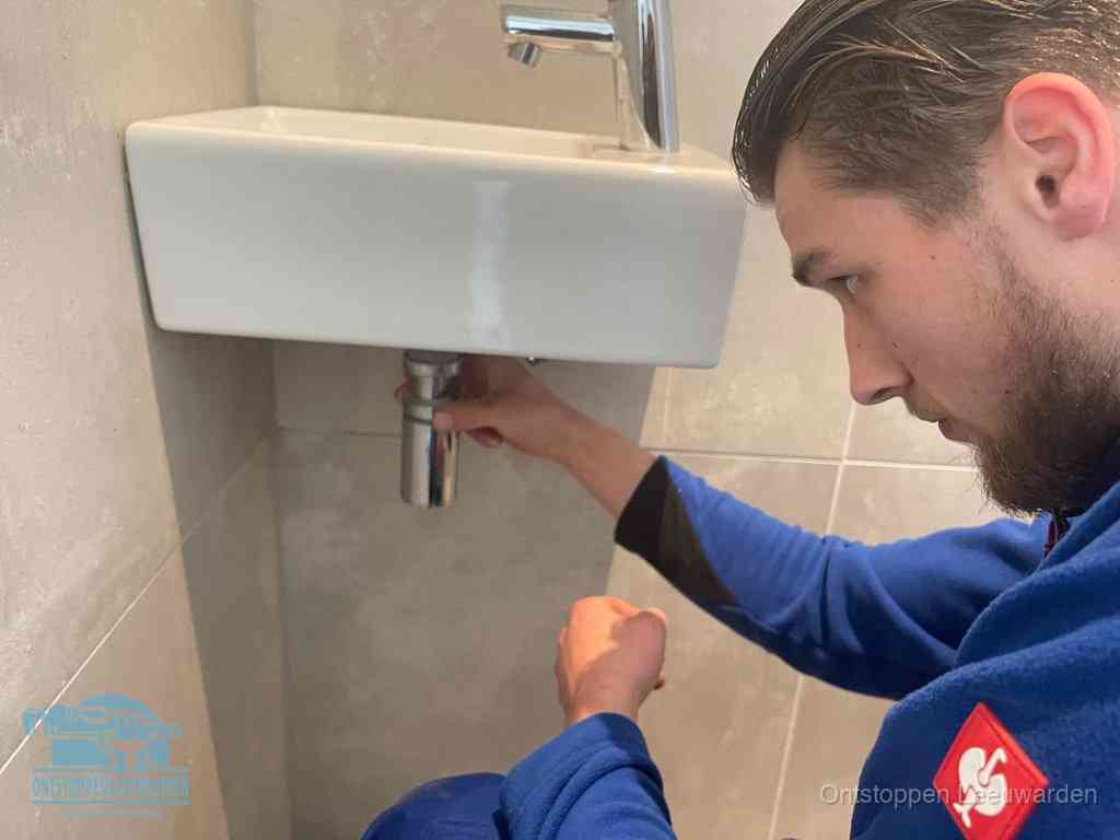 Een monteur draait met de hand de zwanenhalssifon onder een fonteinwasbak in de toiletruimte los om de bron van rioollucht in Leeuwarden te vinden