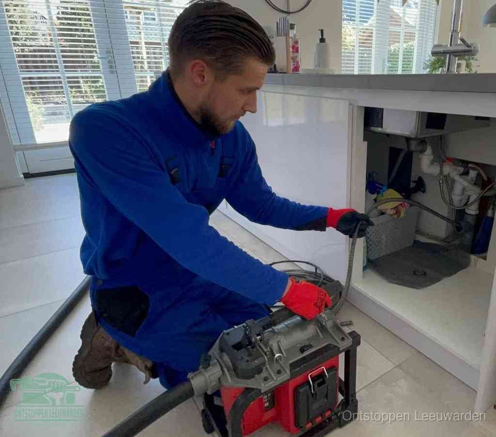 Een monteur met rode handschoenen knielt in een keuken en voert een elektrische ontstoppingsveer in tijdens het professioneel afvoer ontstoppen in Leeuwarden